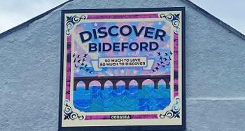 A colorful mural on the side of a building featuring the words 'Discover Bideford' in bold letters. Below, a smaller text reads 'So much to love, so much to discover.' The artwork shows a stylized bridge over blue waves with birds flying in the background