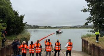Photo of multiple people in orange high vis, being given a talk in front of the river.