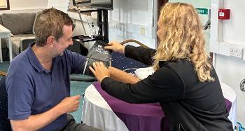 A blonde lady checking the blood pressure of a man by placing the blood pressure cuff on his arm. They are sat down at a table opposite each other. 