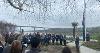 A group gathers outside Riverbank House beside the River Torridge for a Commonwealth Flag‑raising event. The crowd forms a semicircle near a flagpole while the flag is raised. The Torridge Bridge spans the river in the background under cloudy skies.